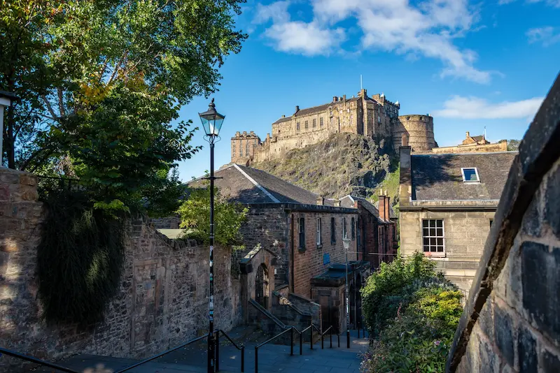 Calle de escaleras con farolas en medio que bajan mientras el castillo de Edimburgo se ve al fondo en lo alto de una colina