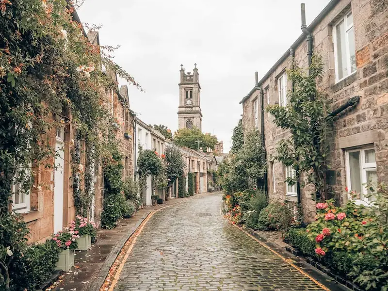 Es una calle de piedra con muchos arbolitos pegados a los muros de piedra y casas pequeñitas. Al fondo, por encima de las casas, se ve una gran torre con un reloj