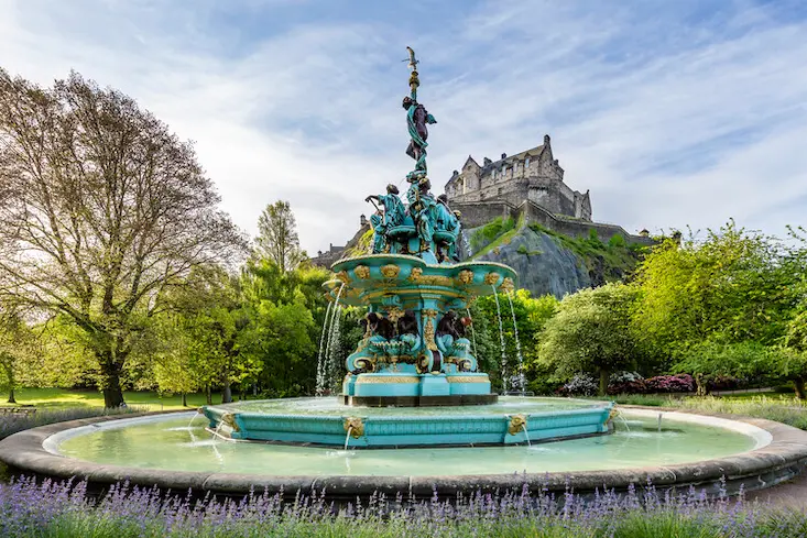 Una fuente en unos jardines de Edimburgo con el castillo de Edimburgo en la colina al fondo. La fuente es grande con 3 niveles y varias figuras