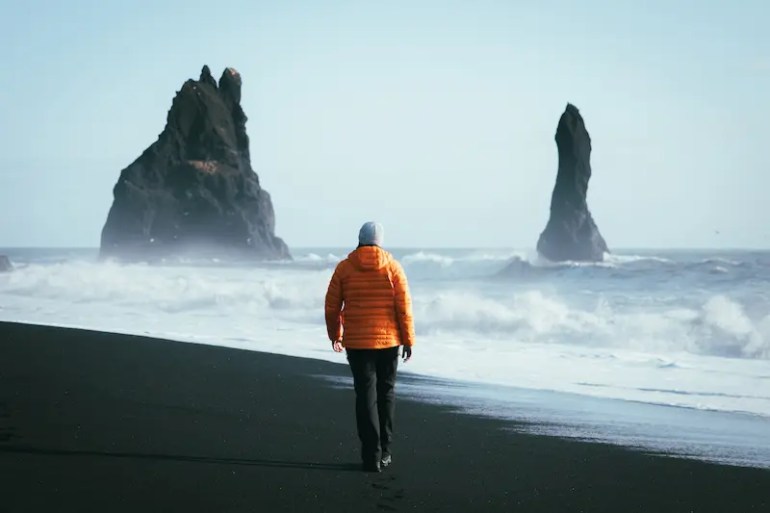 BSB· Un chico anda dirección al mar en una playa de arena negra en Islandia. En el horizonte dos grandes rocas emergen del mar. Un mar lleno de olas y bruma marina