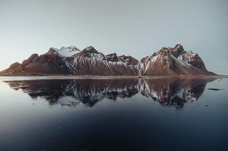 Vestra3 Una cordillera montañosa marrón, llena de nieve en sus picos y sus pies un lago con el agua tranquila. La cordillera se refleja perfectamente en el agua del lago como si fuera un espejo